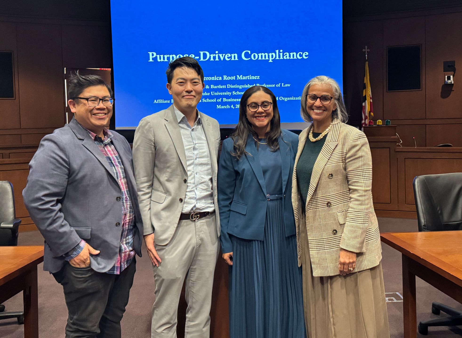 Four people in professional attire smiling in the Ceremonial Courtroom