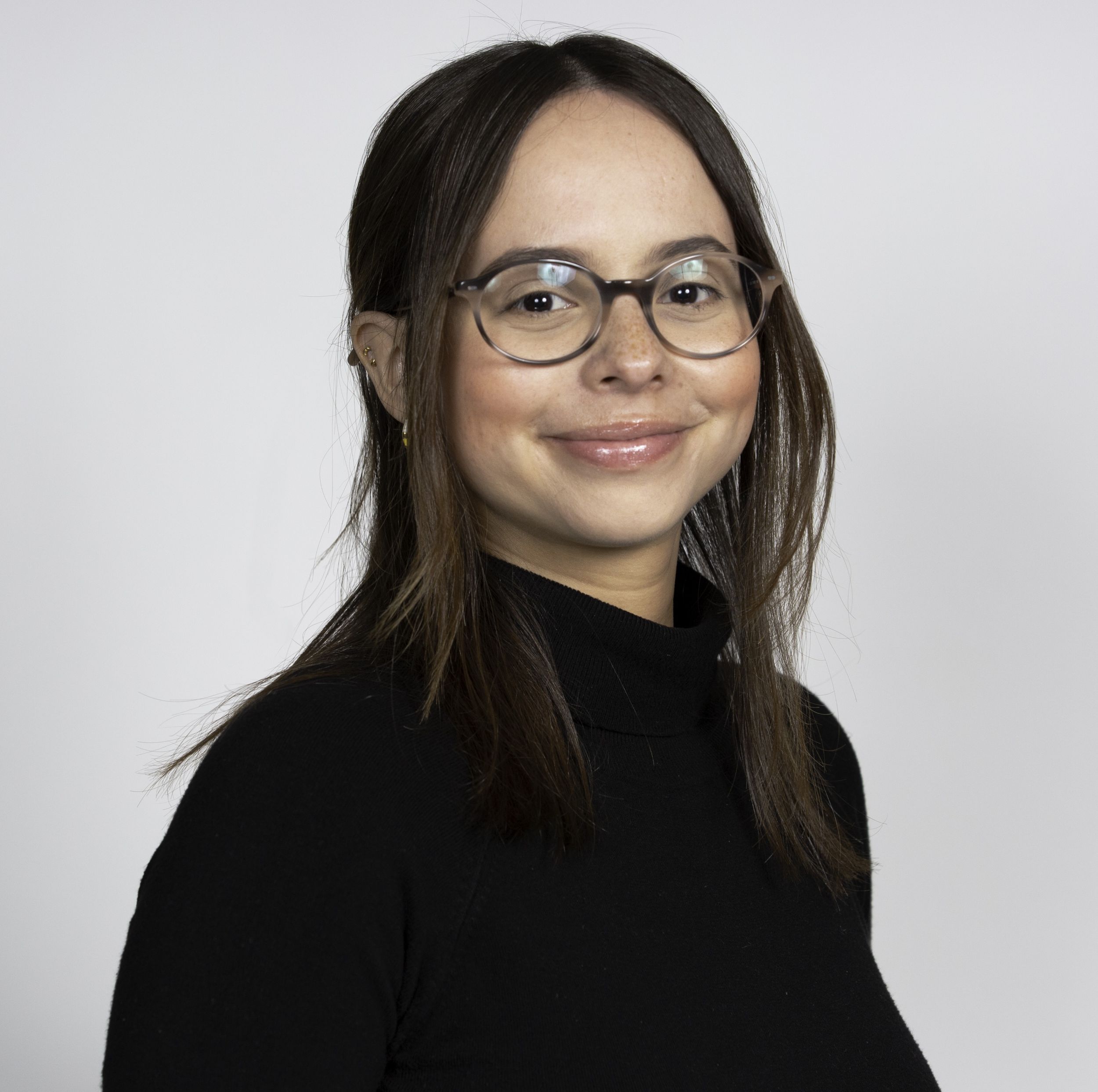 Daniela Benites, with shoulder length brown hair and wearing glasses, smiles into the camera, wearing a black sweater with a grey background