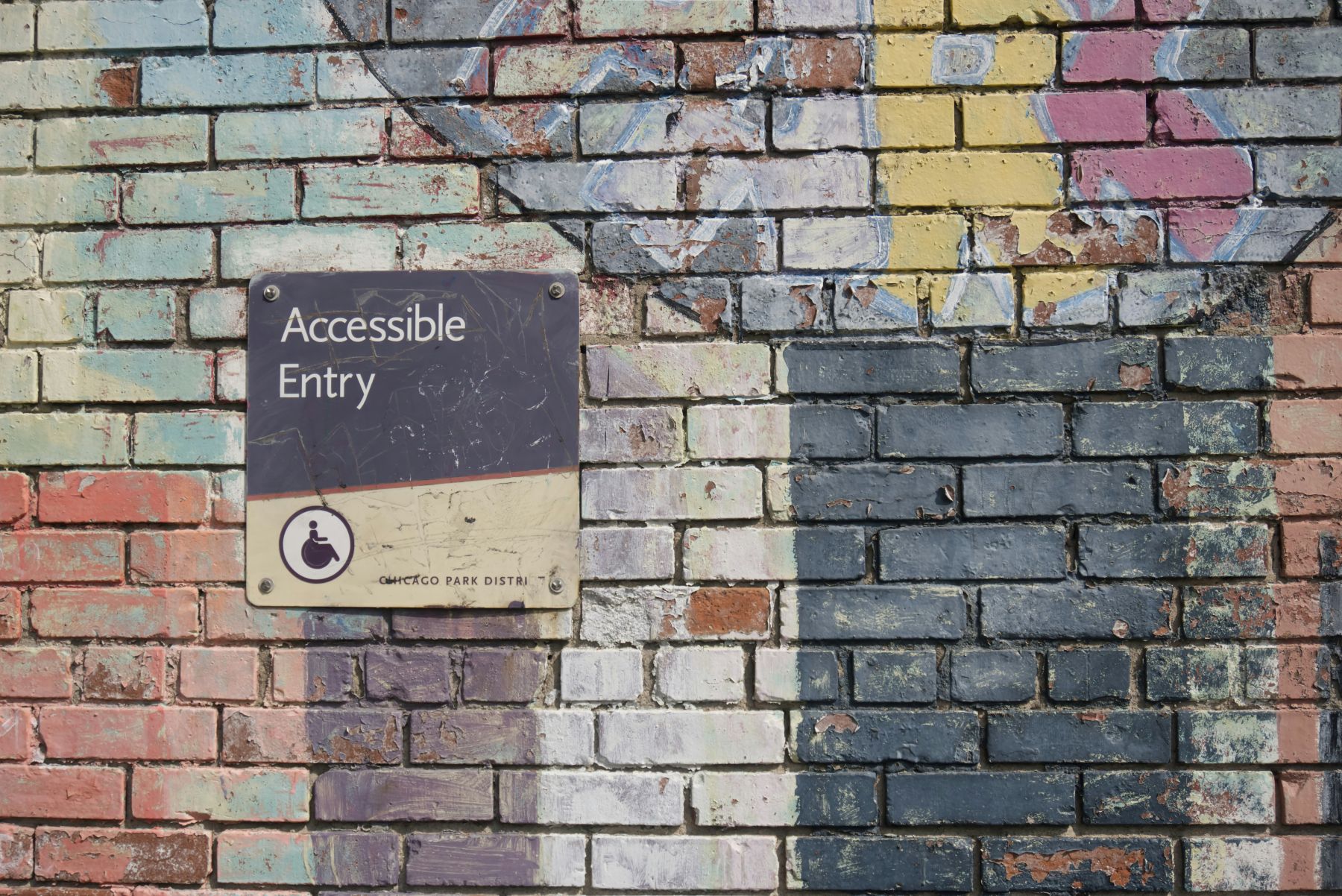 A worn “Accessible Entry” sign with a wheelchair symbol mounted on a brick wall painted with faded, multicolored abstract patterns