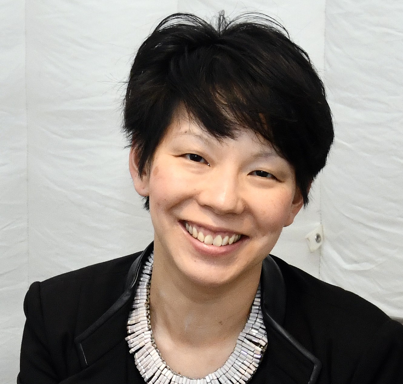 Julie, with short dark hair, looks smilingly directly at the camera, wearing a black blazer over a dark top and a distinctive white beaded necklace.