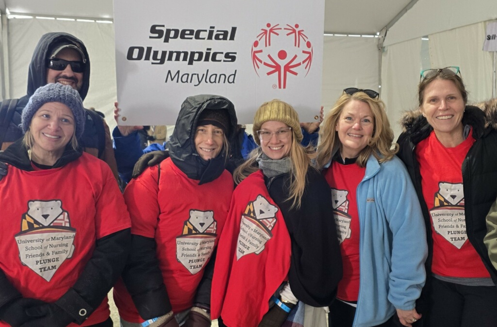 Group of nursing faculty in red University of Maryland School of Nursing Friends & Family Plunge Team shirts stand under a tent, holding a Special Olympics Maryland sign.