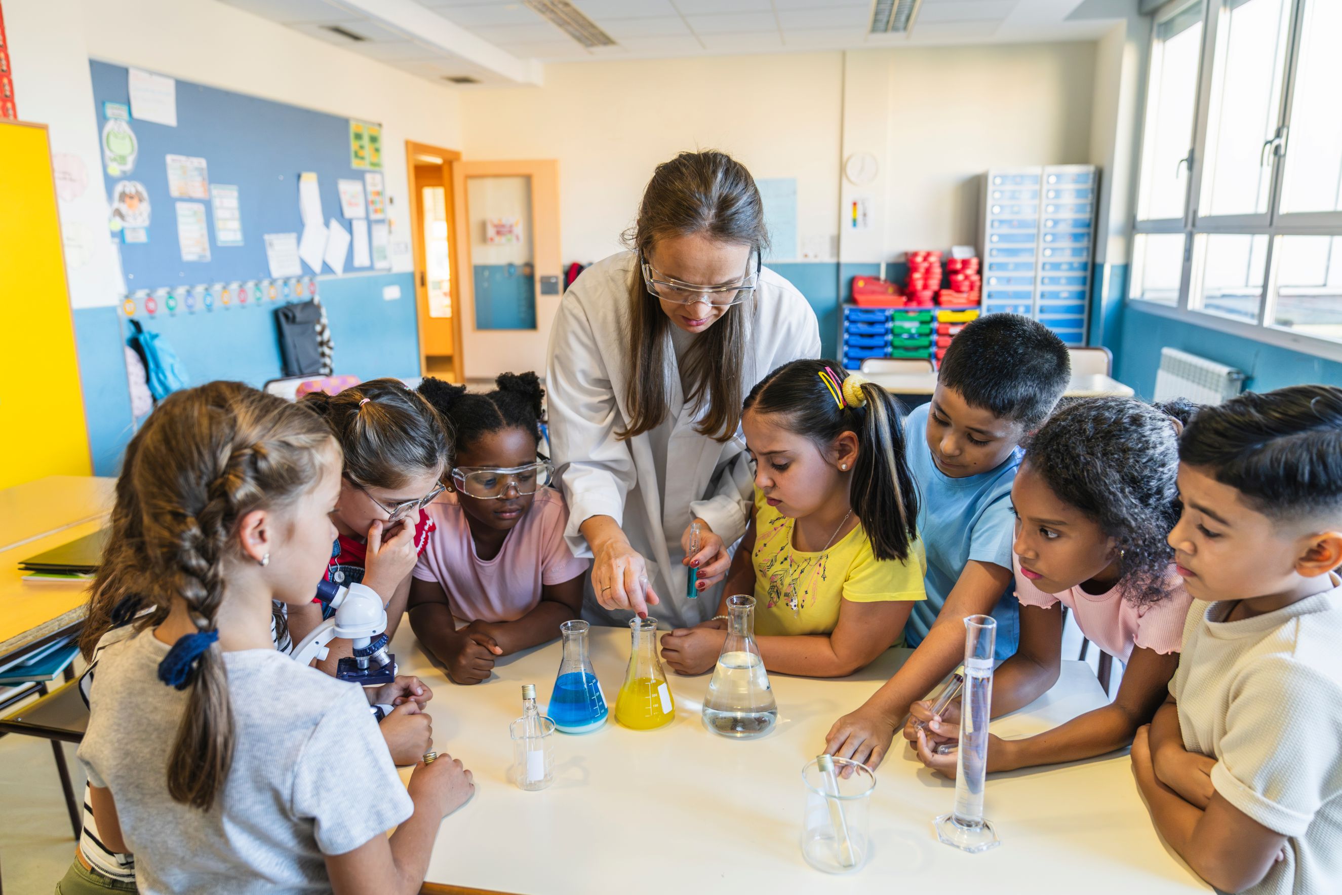 Teacher guides a diverse group of children conducting a science experiment with colorful liquids, laboratory glassware, safety goggles, and a microscope.