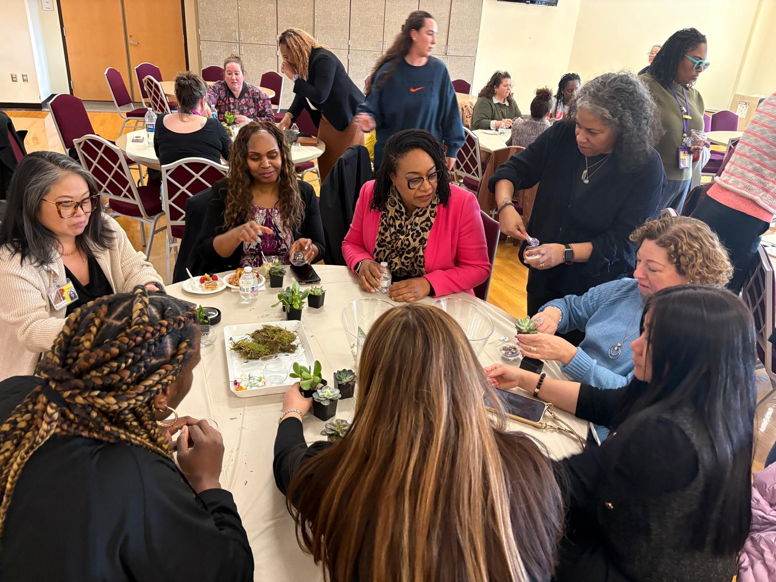 A group of about ten women are seated and standing around a round table in a bright indoor room, engaging in a hands-on activity of creating small succulent terrariums.