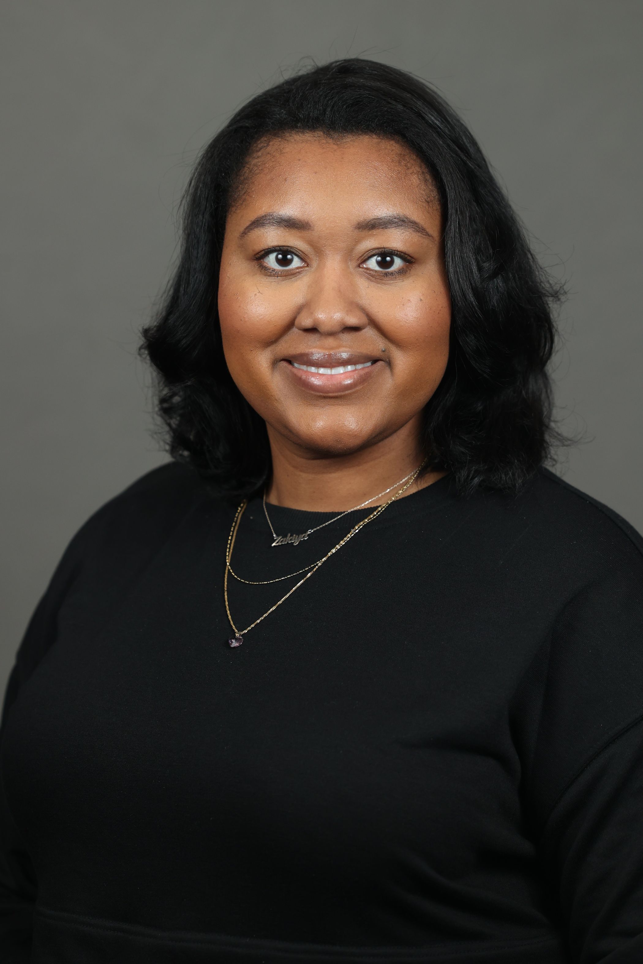 Zakiya Whatley, with shoulder-length black hair wearing a black top and layered necklaces, smiles and faces the camera against a plain gray background.