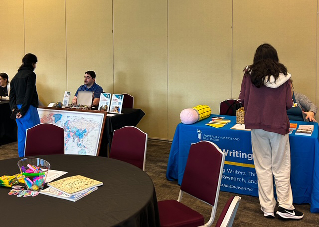 A conference room with two booths. One features maps and a seated person, the other has a blue tablecloth with brochures. People interact casually.