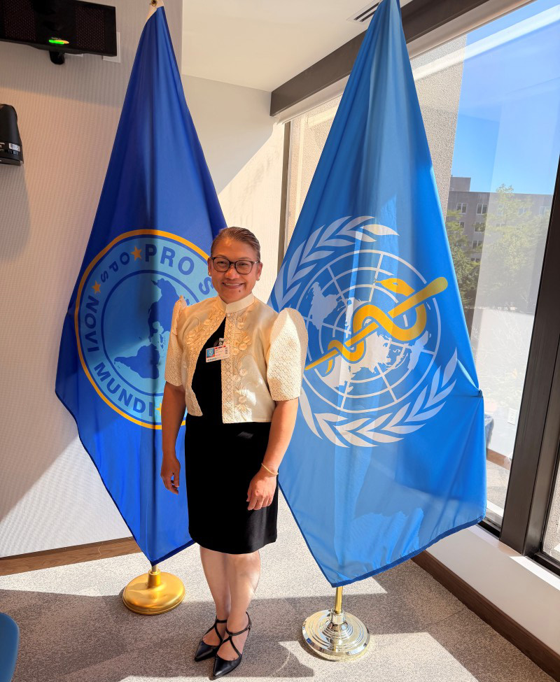 Angelica Fernandez-Dizon stands smiling between two blue flags, one with a globe and snake symbol, in a brightly lit room. The mood is professional and welcoming.