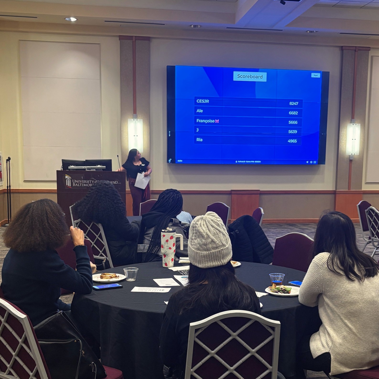 Staff presenting at a podium in a university conference room while a scoreboard is displayed on a large screen. Attendees sit at round tables with food, watching the interactive session.