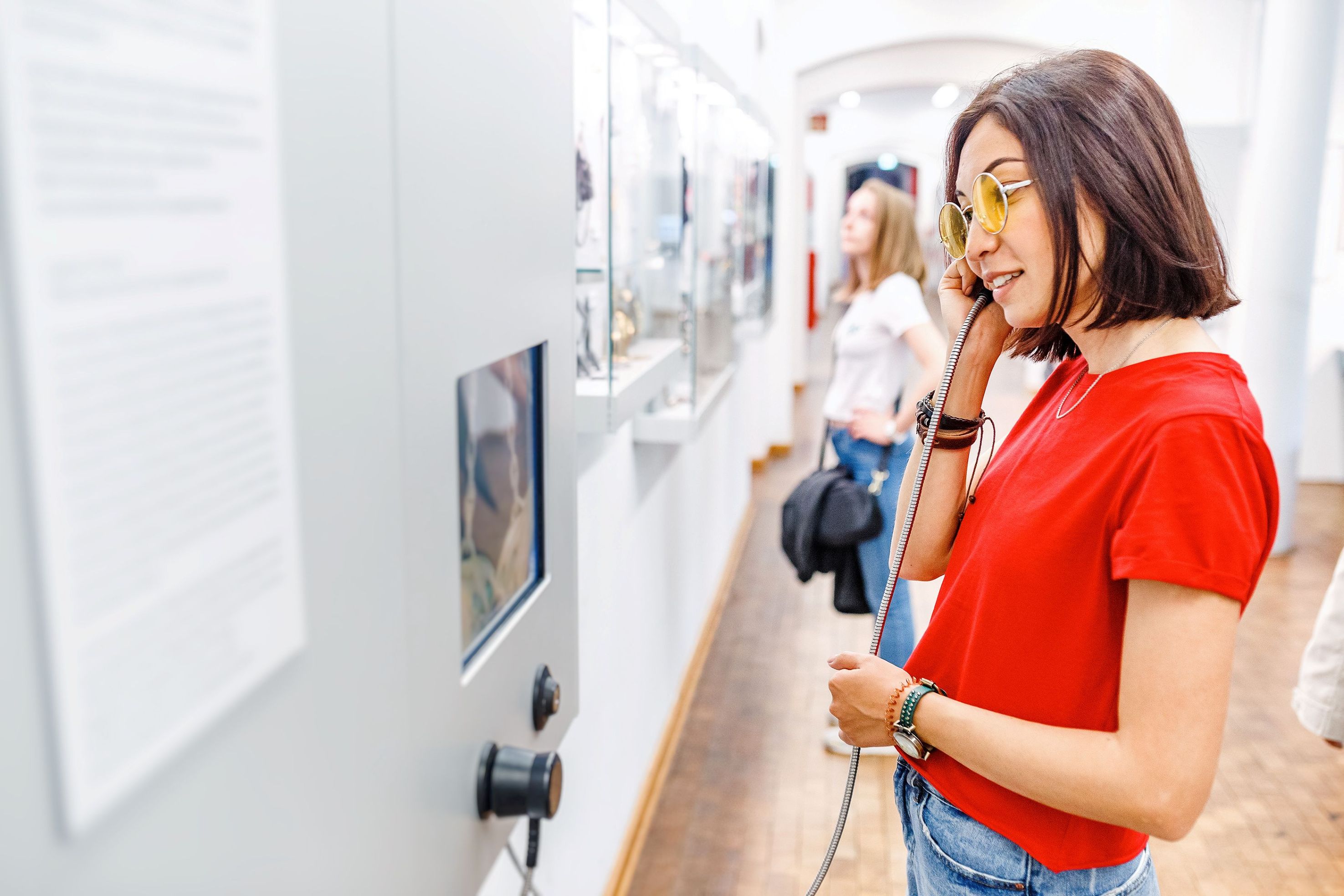 Museum visitor in a red shirt listens to an audio guide while viewing a display case of historical objects. Another visitor explores exhibits farther down the gallery.