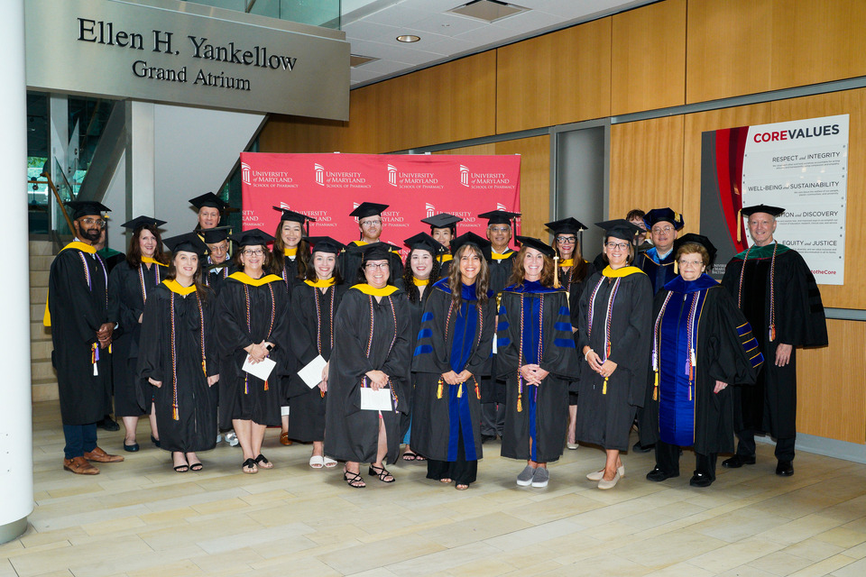 Group photo of palliative care graduates and program director in graduation caps and gowns in front of a red School of Pharmacy banner