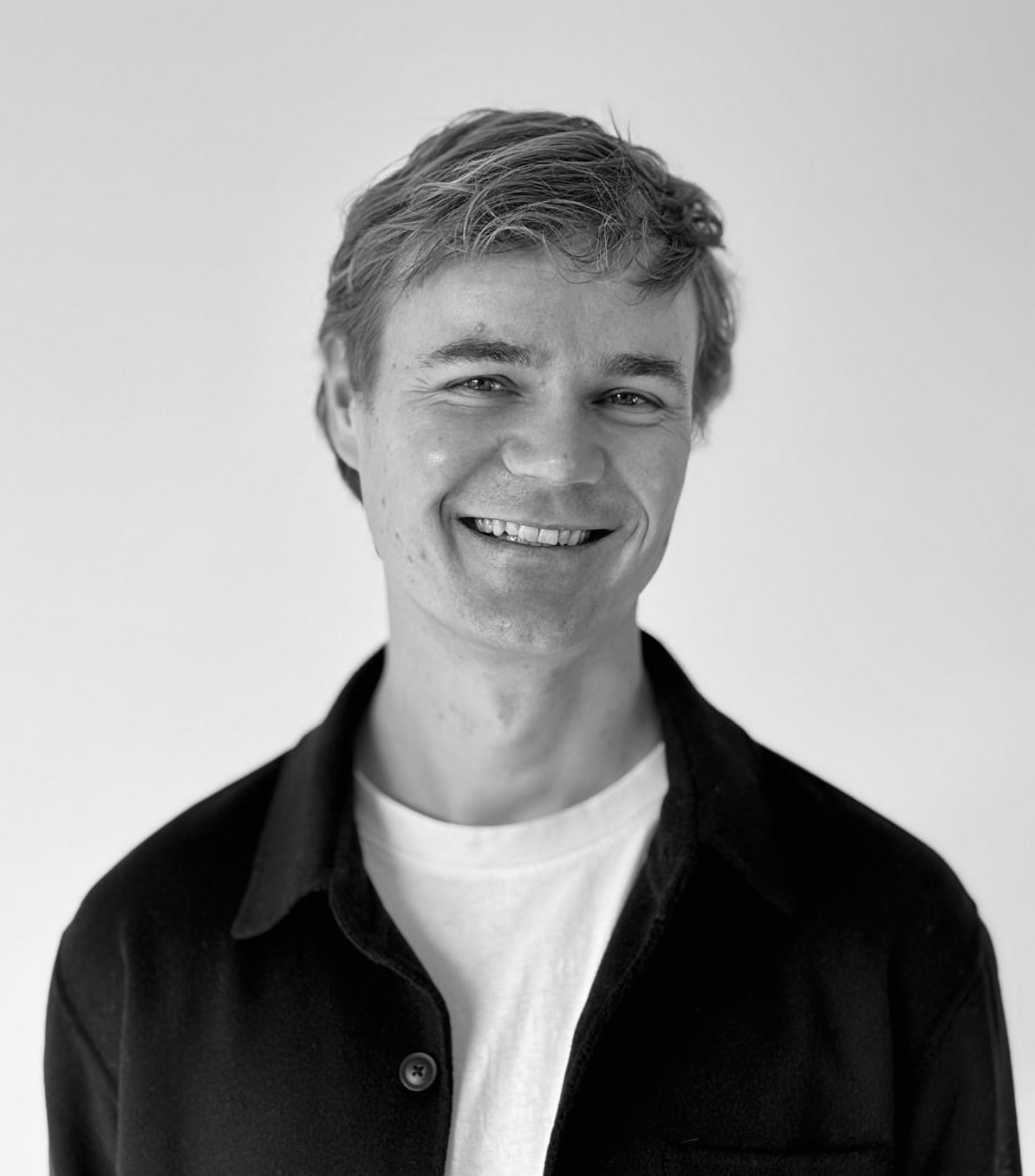 Black-and-white portrait Lucas Kreuzer, smiling, with short, tousled hair, wearing a dark collared jacket over a light T-shirt against a plain, light background.