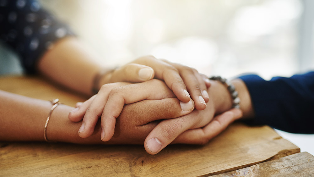 Two pairs of hands gently clasp each other on a wooden table, conveying a sense of comfort and support. The background is softly blurred and sunlit.