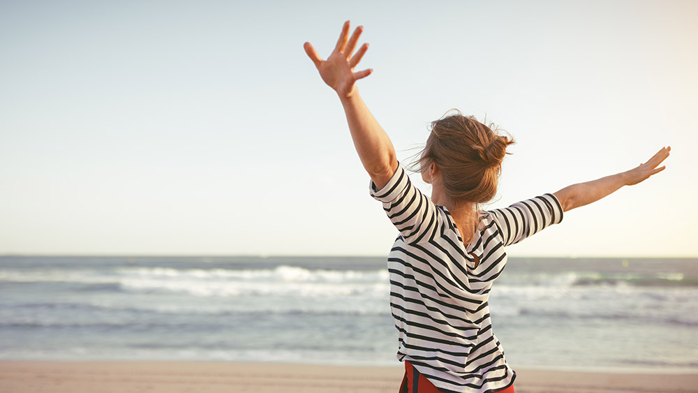 A woman in a striped shirt stands on a beach with arms outstretched, embracing the sea breeze. The setting sun casts a warm glow, evoking freedom and joy.
