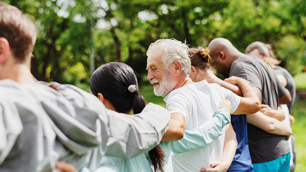A diverse group of adults stands outdoors in a circle with their arms around one another, representing community, connection, and mutual support.