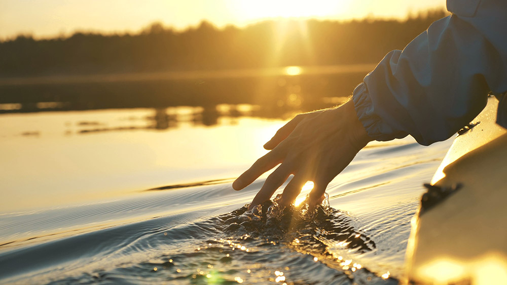 A hand gently skims the water's surface from a boat at sunset, creating ripples. The golden light reflects off the tranquil lake, conveying serenity.
