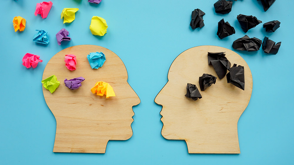 Two wooden silhouettes of heads on a blue background, one filled with colorful paper balls, the other with black ones, symbolizing contrasting thoughts.