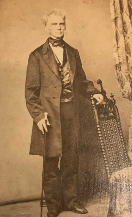 Sepia photograph of an older man leaning against a chair, the man is wearing a long coat, bow tie, and vest.