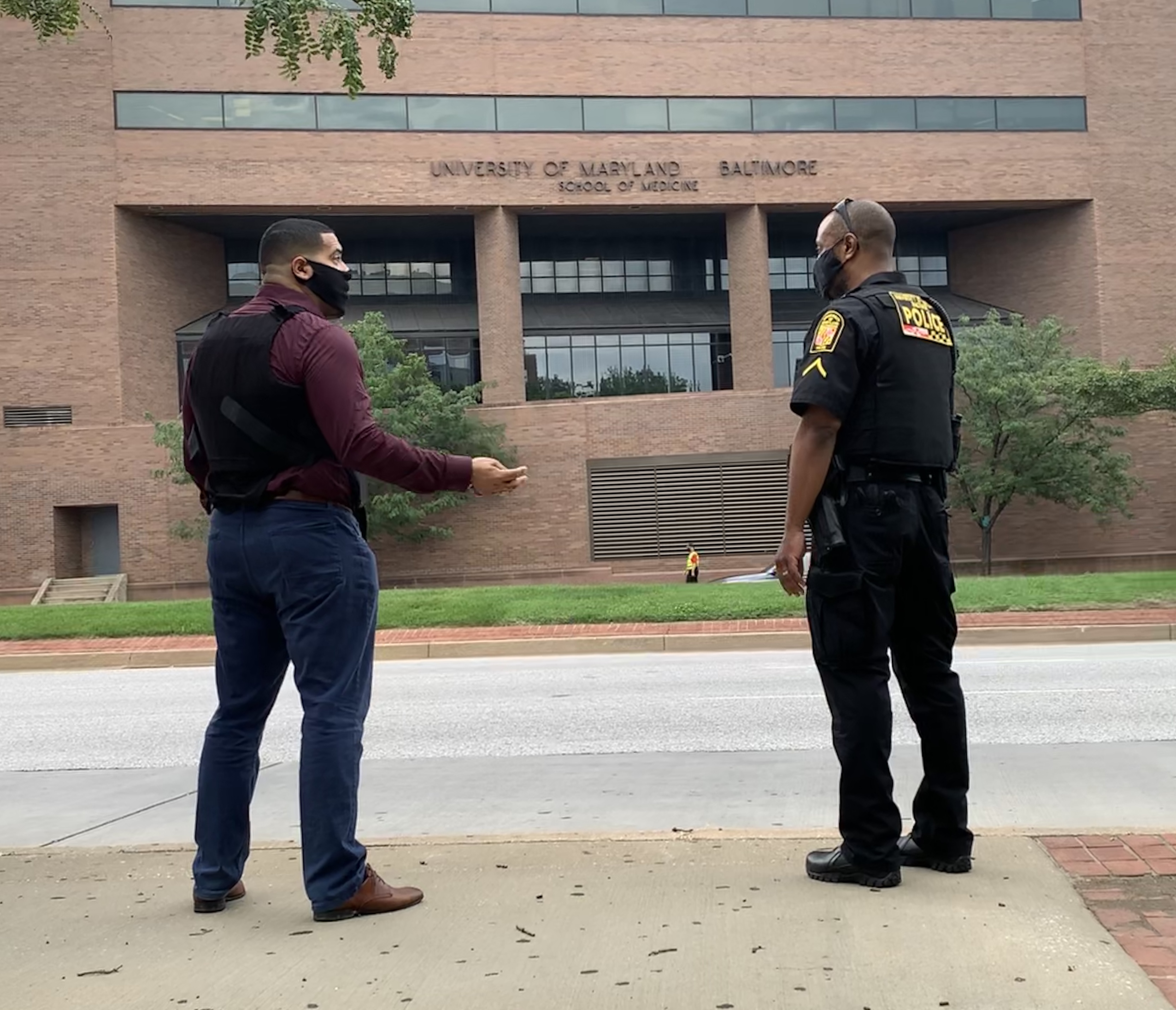 Jonathan Alfaro and Pfc. Yale Partlow stand socially distanced wearing masks, facing each other and talking. The University of Maryland, Baltimore School of Medicine building is shown behind them.