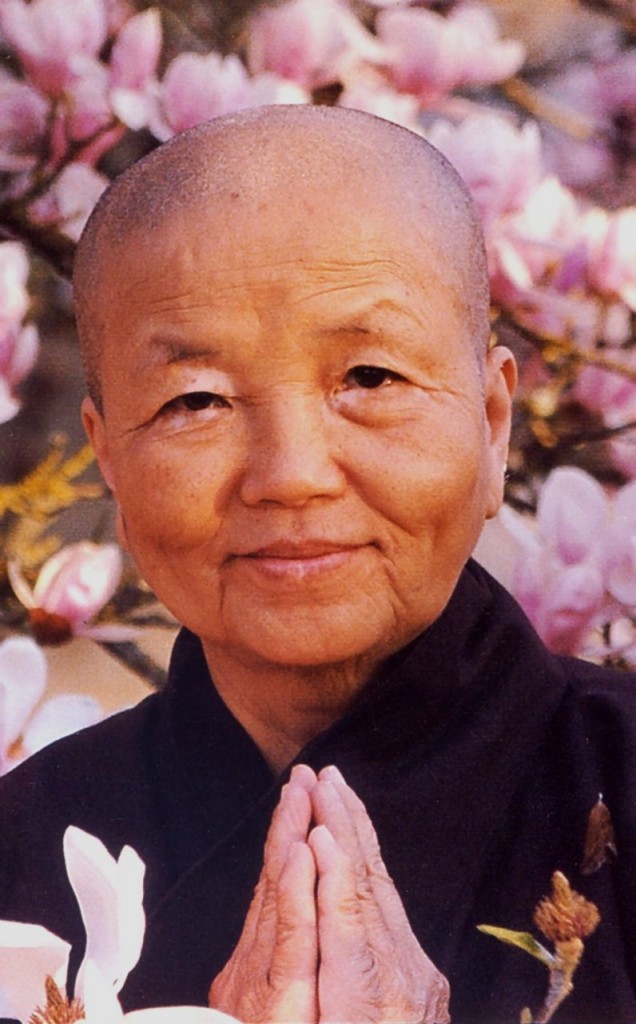 Image of Sister Chan Khong with the palm of her hands together at her chest, standing in front of pink flowers